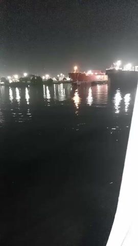 The video captures a nighttime scene at a port or a large waterway, showing several large ships docked or anchored. The scene is dark, illuminated primarily by numerous bright artificial lights originating from the ships and the distant shore, which create prominent, shimmering reflections on the water's surface.

The camera slowly pans from left to right, revealing different sections of the vessels. Initially, a reddish-brown ship is visible on the left side of the frame. As the camera pans, a larger, darker-hulled ship comes into view. This ship appears to be a tanker or cargo vessel, with a long, flat deck and a superstructure towards the stern. The name "CABO DE HORNOS" is clearly visible in white letters on its bow.

Throughout the pan, bright lights are seen on the decks and superstructures of the ships, as well as on structures along the shoreline in the background. These lights cast long, distorted reflections on the rippling dark water, creating a dynamic interplay of light and shadow. Towards the end of the video, the stern of the "CABO DE HORNOS" ship is visible, along with what appears to be a smaller vessel or tugboat positioned near its rear. The video seems to be filmed from a moving boat, as suggested by the slow, continuous panning motion and the white railing visible on the far right of the frame. The overall atmosphere is serene, with the dark water contrasting with the glowing lights.