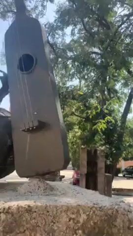 The video shows an outdoor sculpture of several musical instruments. The largest part of the sculpture is a tall, angular guitar with a soundhole and four strings. Beside it is a spherical object with a crown-like structure on top, and behind the sphere, a long, cylindrical instrument appears to be leaning against the guitar. The sculpture is set on a stone base surrounded by what looks like dirt and small stones. The background includes trees, a clear blue sky, and part of a building in the distance. The video pans upward, revealing more of the sculpture against the sky.