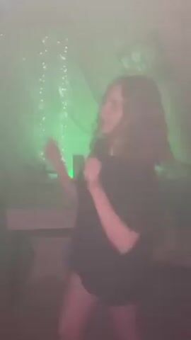 A woman with curly brown hair, wearing a black t-shirt, dances in a room filled with a fog-like haze. The room is illuminated by different colored lights, including green, blue, and pink. In the background, there are string lights and a few blurred objects, one possibly a table. The woman is facing the camera and singing along to the music. Her body language is animated as she moves to the beat.