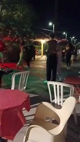 At night outdoors, several couples are dancing. In the foreground, there are white plastic chairs and red-clothed tables. In the background, there are palm trees, lights, and more people. Two people are dancing close together in the center of the frame. To the left, another couple is dancing. To the right, a man in a white shirt and dark pants is dancing. A stage area with green lighting is visible on the far left, with two people standing on it. The overall atmosphere appears to be festive.