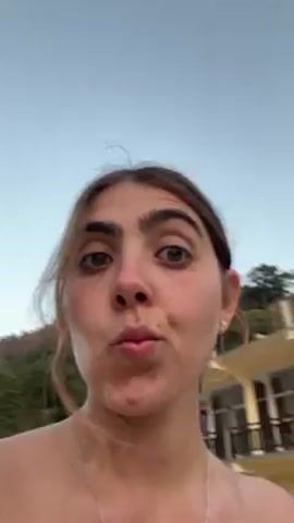 A young woman, fair-skinned with dark eyebrows and brown hair pulled back, is recording a selfie video. She is wearing a thin silver necklace and small earrings. Her top is out of frame, but it appears to be patterned. She makes various facial expressions, including pursing her lips and sticking out her tongue slightly. Initially, the background shows a building with a red tile roof and lush greenery. The camera then pans downwards to show a gray concrete surface and some outdoor furniture. Finally, the background shifts to reveal a bridge spanning a body of water, with mountains visible in the distance under a clear sky.