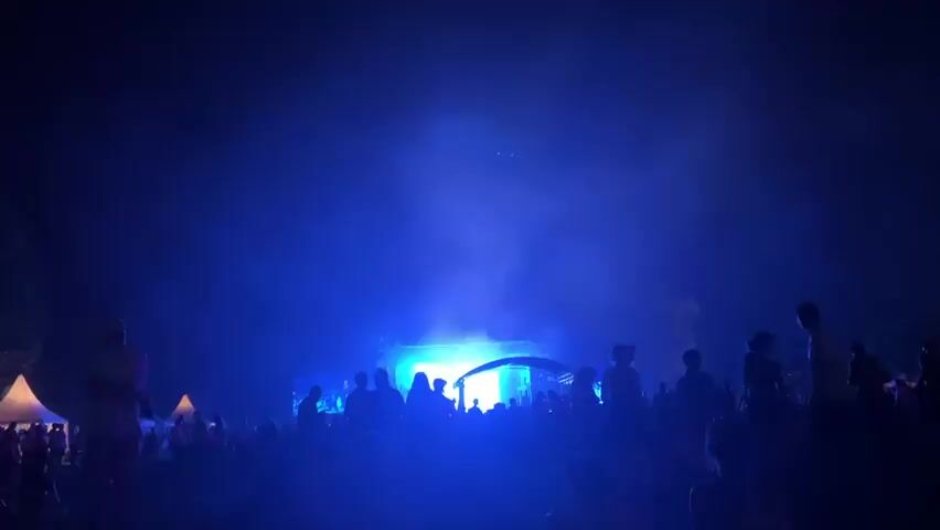 A large crowd of silhouetted people stand in front of a brightly lit stage at night. The stage is illuminated by vibrant blue lights, creating a smoky effect in the air. Some people in the crowd appear to be dancing or moving to music. The scene suggests an outdoor concert or festival event.