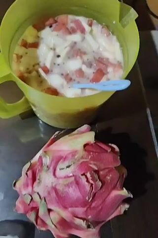 On a metallic surface, there is a yellow pitcher filled with a mixture of chopped fruits in a liquid. There appears to be watermelon, white translucent pieces, and some dark seeds in the mix. A blue plastic spoon rests inside the pitcher. Next to the pitcher lies a whole dragon fruit with a pink rind. The camera slightly adjusts its angle and zooms in a bit. In the background, there's a small, empty glass.