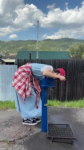The video shows a woman in a blue dress, white t-shirt, red cap, and black-and-white shoes, standing next to a blue water pump on a street. She is bending over to drink water from the pump, then straightening up and smiling at the camera. She also has a red and white plaid shirt tied around her waist. The background includes a wooden fence, a house with a green roof, and a mountain range. The sky is blue with scattered clouds. The ground is paved, with a manhole cover and a metal grate visible.