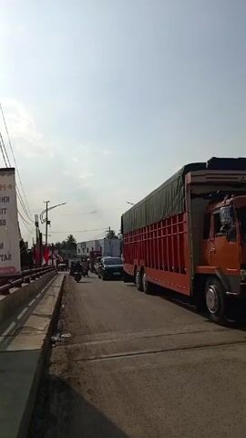 The video shows a view of a busy street during the day. On the right side of the road, a large, red truck with a covered bed is stopped. To the left, there is a narrow sidewalk, a road barrier, and a banner with text written on it. Cars and motorcycles are seen in the distance, moving along the road. The road surface is dry and appears to be in average condition. Power lines and streetlights are visible overhead. The sky is mostly clear with some clouds.