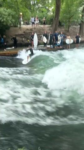 This video shows a surfer in the middle of a river, riding a standing wave. The surfer is wearing a black wetsuit and is standing on a white surfboard. The wave is large and foamy, and the river water is green. There are several people watching the surfer from the riverbank. The riverbank is lined with trees and there is a dirt path behind the spectators. Some of the spectators are holding surfboards. The video is shot from a distance, and the focus is on the surfer and the wave. The footage appears to be taken during the daytime, as there is natural light.