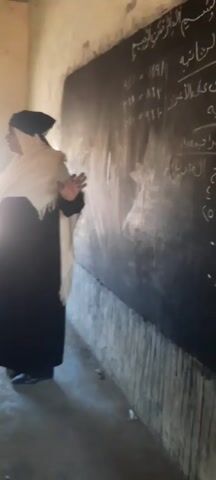 The video shows the interior of a classroom with plaster walls and a barred window, illuminated by natural light. A teacher in a black dress and a white shawl stands beside a black chalkboard with Arabic script written on it, in front of rows of students seated at desks. The students wear green and white uniforms. Some have books and notebooks on their desks. A long black stick is seen on a desk near the teacher.