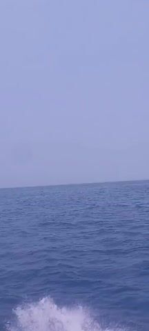 The video shows a body of water, likely the ocean, with a hazy sky. The camera appears to be on a moving boat, as evidenced by the water splashing at the bottom right of the frame. In the distance, two boats are visible moving across the water. The first boat is small and appears to be farther away. The second boat is larger and closer, leaving a white wake behind it as it speeds across the water. The overall scene suggests a bright, possibly overcast day at sea.
