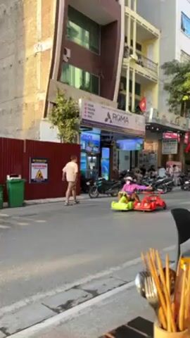 Here's a description of the video:

The video shows a person riding a connected pair of small, plastic toy cars on a city street. One car is red, and the other is green. The person riding them is wearing a purple jacket and white face mask. They appear to be having fun, with their arms spread out. 

The scene is set on a paved road in an urban area, with buildings on either side. There's a red corrugated metal fence visible along the side of the road. A motorbike passes the person on the toy cars. In the foreground, on the right, is a wooden utensil holder, suggesting the video is taken from a restaurant or eating area.