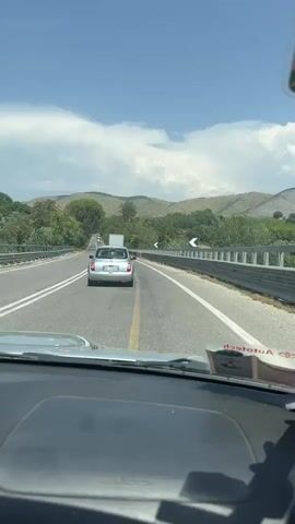 The video is taken from inside a car, looking out the windshield. The car is on a highway with two lanes in each direction, separated by a yellow line. There are other cars and trucks on the road ahead. The landscape is hilly and green, with trees and vegetation on either side of the road. The sky is blue with some white clouds. There is a white car directly in front of the filming car, followed by a truck. The filming car has a parking permit and a sticker on the dashboard. The timestamp at the beginning of the video is 00:00.