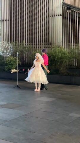 A person in a pink shirt and dark pants styles a person wearing a short white dress with pink accents. Both are standing in a public plaza setting. The person in the dress has a headpiece with a veil. There is a phone on a stand pointed at them. Behind them is a tall, rectangular building. Shrubbery is on either side of them. There is a backpack, water bottle, and plush toy visible on the ground. They begin to sway to music.