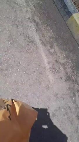 The video shows a person's feet walking on a wooden walkway. The walkway is adjacent to a paved area with a yellow and gray curb. In the background, a blue surface, possibly water, is visible. The camera is held at a low angle, focusing on the ground and the movement of the feet.