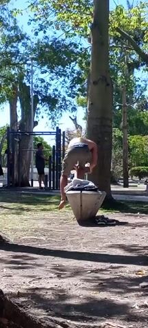 The video shows a man shirtless and wearing shorts bending over a trash receptacle in a park on a sunny day. The receptacle is light gray in color and inverted cone-shaped. The man is facing away from the camera. Behind the man are several trees, a metal structure that appears to be exercise equipment, and another person standing near the structure. There are shoes lying on the ground next to the trash receptacle. The ground is dirt with grass. There are also trees on the left and right sides of the shot. The sky is blue with some clouds.