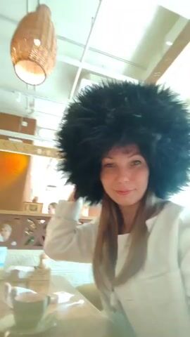 The video opens with a close-up shot of a woman wearing a large, fluffy black hat. She is smiling and looking at the camera. She is seated at a table in what appears to be a restaurant or cafe. Behind her, a wicker lampshade hangs from the ceiling, and there are other patrons in the background. The video then cuts to a young woman wearing a large, fluffy white hat and glasses, sitting at the same table. She is also smiling. The shot pans slightly to show another person on the right side of the frame, also wearing a fluffy hat, and a table with drinks and food items. The scene appears to be indoors with natural light coming from above.