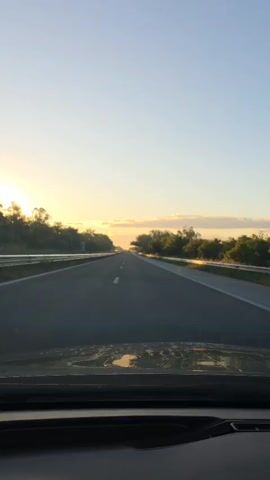 The view from inside a vehicle driving down a highway shows the road stretching ahead under a clear, blue sky tinged with the orange glow of the rising sun. Trees line both sides of the highway, with metal guardrails on the shoulders. The road surface appears slightly wet or speckled with moisture, reflecting light. There is a white lane marker dividing the lanes.  The dashboard of the vehicle is visible in the foreground. The video captures a sense of movement as the vehicle progresses along the highway.