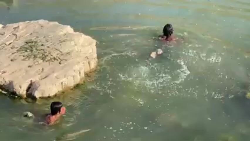 Here's a detailed description of the video:

Two young individuals are seen swimming in a body of water. The water appears somewhat murky, with a greenish tint. To the left of the frame, there is a light-colored, rocky outcrop with some green vegetation growing on top. The individual closer to the camera is swimming with their head above water. The other individual is further away and submerges, causing a splash. The overall environment appears to be outdoors, possibly a natural pool or pond.