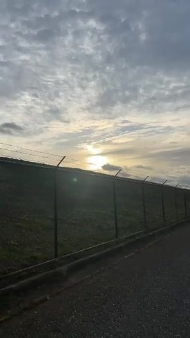 The video shows a chain-link fence with barbed wire running along a grassy embankment next to an asphalt road. The sun is visible in the sky, partially obscured by clouds, and the light suggests it may be sunrise or sunset. As the camera pans down the road, three figures can be seen in the distance, appearing to be walking. The overall environment is outdoors, with a clear sky and vegetation.
