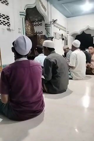 Several men are seated on the floor of a room, likely a mosque or religious space. They are all facing the same direction, toward an ornate structure, likely a mihrab. Each man wears a white or light-colored religious cap. Some of the men are reading from books or papers they hold in their hands. The room is well-lit, with a white tile floor and light-colored walls. The ceiling appears to be painted a light blue. There is a small shelf with books behind some of the men and a fan attached to the wall. One man, in the foreground, is wearing a purple shirt. The overall atmosphere seems to be one of quiet contemplation or prayer.