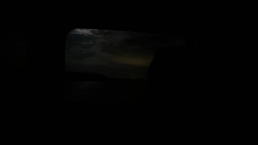 The video shows a dim view through a window, possibly from inside a vehicle, overlooking a body of water at night. The moon is visible through the clouds, casting a faint reflection on the water. There is a dark landmass on the other side of the water. The camera moves slightly, and there is music playing in the background.