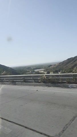 The video depicts a roadside view from a moving vehicle. A gray concrete barrier runs along the edge of the highway, separating the road from a valley below. The valley is filled with vegetation and what appears to be a dry riverbed or sandy terrain. The sky is clear and light blue, indicating a sunny day.