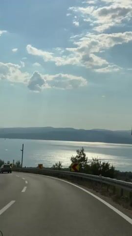 The video shows a POV shot from a car driving on a road that curves to the left. The road has white lane markings and a metal guardrail on the right side. There are yellow and red warning signs indicating sharp curves ahead. Beyond the road and guardrail, there is a body of water, likely the sea, with mountains in the distance. The sky is blue with scattered clouds. There are trees and vegetation along the side of the road.