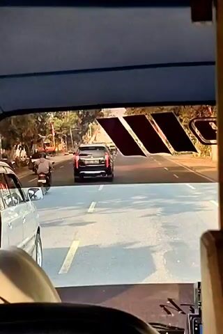 The video shows a view from inside a vehicle, looking out through the windshield. The perspective is from the driver's or passenger's seat, with a portion of the vehicle's dashboard and steering wheel visible in the foreground. The road ahead is paved and marked with white lane lines. A black SUV is traveling in the same direction as the viewer's vehicle, in the lane to the right. To the left of the SUV, a person is riding a motorcycle. Further down the road, several other vehicles, including cars and trucks, are visible, as well as more motorcycles and pedestrians on the sides of the road. The environment appears to be a sunny day in a somewhat urban or suburban area, with trees and buildings lining the road. There is a large logo or graphic with three dark, slanted rectangular shapes on the top edge of the windshield. The video captures the normal flow of traffic.