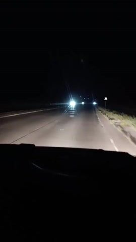 The video shows a first-person perspective of driving on a road at night. The headlights of the vehicle illuminate the wet road ahead, with other vehicles visible in the distance. The sky is dark, and there is a triangular road sign on the right side of the road. The dashboard of the car is visible in the foreground.