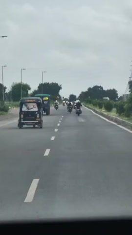The video shows a view from inside a vehicle moving on a paved road. In the foreground, a black auto-rickshaw is visible, decorated with images on its rear. Several motorcycles are traveling ahead of the auto-rickshaw in the same lane. On the left side of the road, there's a blue truck and sparse vegetation. The right side of the road is bordered by a concrete barrier and more trees. The sky is overcast. The overall scene depicts traffic on a highway.