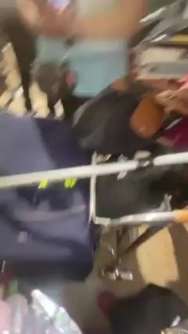 The video shows a person holding a phone camera while moving through an airport. Two young girls, wearing matching shirts and standing next to each other, are briefly in the shot. The camera moves to show other people with luggage. The video ends with the person holding the camera saying "Mommy poo."