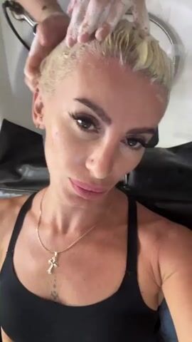 A woman with short, blonde hair and heavy makeup is lying back in a salon chair, with her head tilted back over a sink. Her hair is being washed by another person whose hands are visible, lathering shampoo into her hair. The woman is wearing a black tank top and a gold necklace with a cross pendant. The environment appears to be a hair salon.