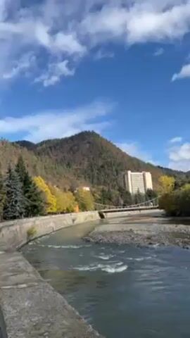 The video presents a scenic view of a river flowing through a mountainous region. The river runs swiftly, with visible ripples and small waves on its surface. A concrete embankment lines one side of the river, suggesting a managed waterway. A suspension bridge spans the river, connecting the embankment to the opposite bank. On the far bank, lush greenery covers the landscape, with trees displaying autumnal colors. A tall building stands amidst the trees, indicating a possible settlement or resort area. The sky is a clear blue, dotted with fluffy white clouds, enhancing the overall picturesque nature of the scene.