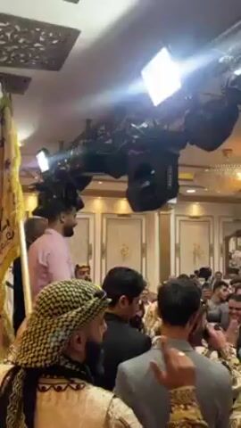 A large group of men are gathered in what appears to be a hall or ballroom, possibly for a celebration or wedding. Many are clapping and smiling, some are dressed in traditional Middle Eastern clothing, including keffiyehs. A man in a pink shirt is being held aloft on someone’s shoulders. Above the crowd, there is a professional lighting rig with spotlights and audio speakers. The room is decorated with ornate molding and gold accents.