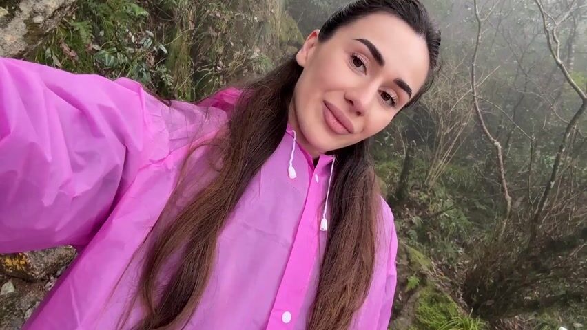 A woman with long brown hair, wearing a pink raincoat, is standing outdoors in a foggy area with rocks and greenery. She is holding the camera and taking a selfie or recording a video. The background is slightly out of focus due to the fog.