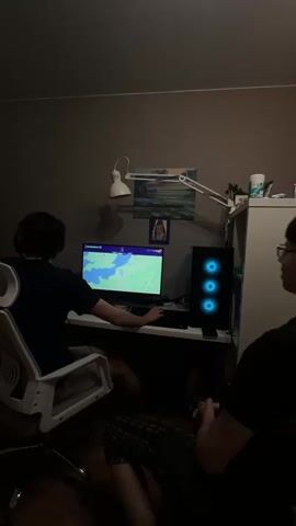 The video shows two people in a dimly lit room. One person is sitting in a white office chair at a desk, playing a video game on a computer. They are wearing headphones and looking at the screen. The game appears to be a racing game. A computer tower with three blue lights is visible to the right of the monitor. Behind the monitor, there is a desk lamp, a picture, and other items on a shelf. The second person is sitting closer to the camera, wearing a black t-shirt and glasses. Their legs are visible, clad in plaid shorts. They are looking towards the computer and appear to be watching the game. The room is otherwise simple, with dark walls and minimal decoration. The camera briefly shifts and rotates throughout the video.