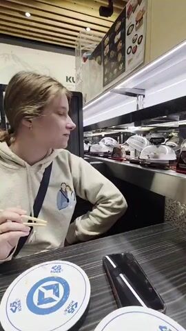 A young woman with blonde hair pulled back, wearing a light-colored hooded sweatshirt with a graphic on the left chest, sits at a table with chopsticks in her right hand. She is looking to her right, off-camera. In front of her on the table are two white circular coasters with blue designs and text, and a black smartphone. Behind her, a conveyor belt system is visible, with plates of food covered in plastic domes moving along the belt. The background appears to be a restaurant setting, possibly a sushi restaurant, with a menu board visible above the conveyor belt. The lighting is bright and artificial.