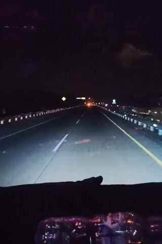 The video shows a first-person view from inside a vehicle driving on a highway at night. The headlights illuminate the road ahead, which has multiple lanes marked by white and yellow lines. Guardrails line both sides of the highway, and various signs and lights are visible in the distance. The interior of the vehicle is dark, with some reflections visible on the windshield, including what appear to be bottles and lights. The overall impression is of a nighttime journey on an open road.
