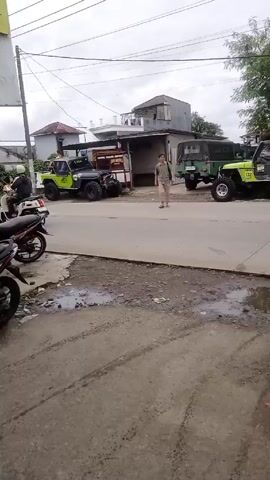 The video shows a man walking on a street.  Several parked motorcycles are on the side of the road.  Behind the man and across the street are two green jeeps.  A sign is visible to the left of the frame.  The sky is overcast.  A building and some foliage are visible in the background.  The street appears to be paved but is stained in places.