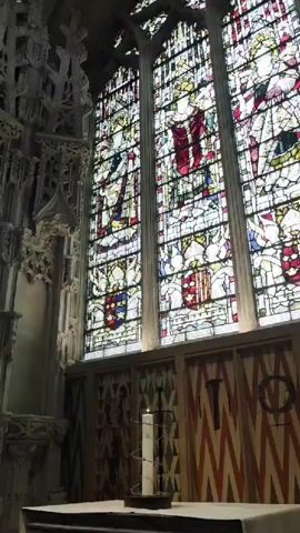 The video pans across the interior of a grand stone building, likely a cathedral or church. The dominant features are large, ornate stained-glass windows and intricate stone carvings. In the foreground, a white altar cloth covers a surface, and on it, a single white candle burns, with a wire coiled around it. The stone carvings are Gothic in style, with elaborate tracery and details. The stained-glass windows depict figures in traditional religious attire, possibly saints or royalty, with coats of arms visible. The lighting suggests it is daytime, with natural light streaming through the windows. The overall atmosphere is one of solemnity and historical grandeur.