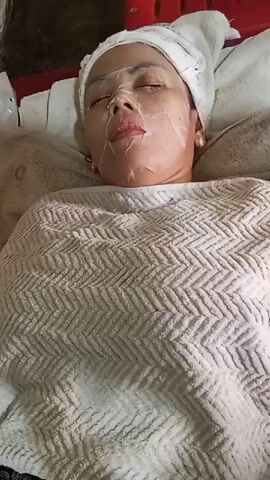 A woman is lying down with her eyes closed. She has a towel wrapped around her head and a sheet draped over her body. Her face is covered with a sheet mask. She is wearing a white towel on her head, and a white patterned towel is covering her chest and upper body. The mask appears to be translucent with creases.