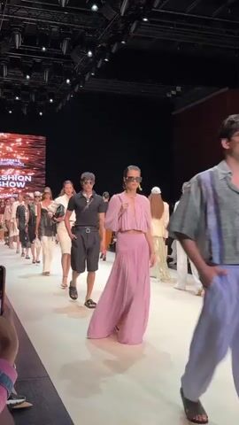 This video shows a fashion show with models walking down a runway. A large screen behind them displays the text "FASHION SHOW". The models are wearing a variety of clothing, including dresses, skirts, shorts, and shirts. Some of the models are also wearing sunglasses and carrying handbags. The runway is white and there are lights on either side of it. The audience is visible in the background, seated on either side of the runway. The video is shot from a low angle, with the camera person's hand visible in the foreground. The atmosphere appears to be energetic and exciting, with the models exuding confidence as they showcase the latest fashion trends.