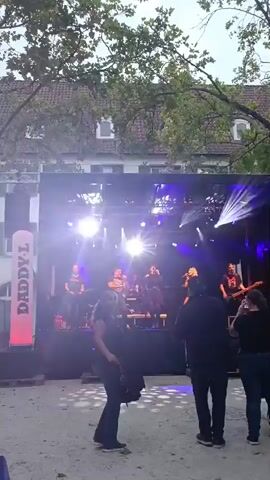 The video shows a band performing on a stage outdoors. The stage is lit with bright purple and white lights. There are several band members on stage, including singers, guitarists, and a drummer. The band is performing for an audience, some of whom are visible in the foreground. There are also trees and buildings in the background. A banner on the left side of the stage reads "DADDY-L". The ground in front of the stage appears to be made of gravel or dirt. The overall atmosphere appears to be that of a live music event.