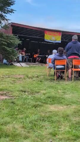 A man in a blue hoodie stands with his back to the camera, facing a band performing on a raised wooden stage. He is wearing khaki shorts. To his right, a woman wearing a grey hoodie sits in an orange chair, also facing the stage. In front of her, another person, likely a woman with dark hair, is visible from the back, also seated in an orange chair. A band of at least five individuals is playing instruments on the stage, which is sheltered by a red-roofed structure with dark siding. A yellow and red flag with an emblem is hung above the band. The stage is set up in an outdoor grassy area with a large tree to the left of the frame. The sky is blue with some clouds. The overall atmosphere appears to be a casual outdoor concert or festival.