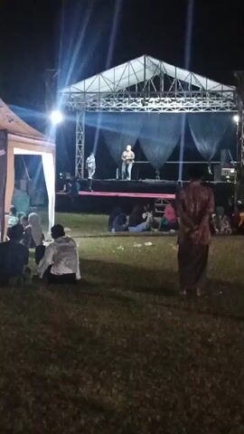 The video shows an outdoor evening event with a stage set up at the back. There are bright lights shining on the stage. On stage, a man is speaking, and another person is standing nearby. A crowd of people is seated on the grass in front of the stage, watching the performance. To the left of the stage, there is a large tent. Inside and around the tent, more people are gathered, some standing and some seated. The ground is covered in grass, and it is dark, indicating it is nighttime. The overall scene suggests a community gathering or festival.