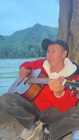 A man with dark hair, wearing a black cap, a red jacket, and dark pants, sits on the ground, leaning against a large tree or rock formation. He is playing an acoustic guitar, strumming with his right hand and fretting with his left, while singing with an open mouth. A wide river with flowing water is visible behind him. Across the river, green trees line the bank, and a large green mountain with a small structure on its peak rises against an overcast sky.