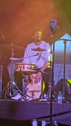 A male musician with glasses and curly hair plays a cajon drum on a stage. He is wearing a white t-shirt and black pants, and white sneakers. In front of him, there is a small snare drum on a stand. There are drum cymbals to his left. A microphone is positioned in front of the cajon, and another microphone is to his right. A camera is mounted on a tripod behind him, angled towards him. The stage is lit with yellow and purple lights, and there are speakers on the right side of the frame. A water bottle is visible on the stage.