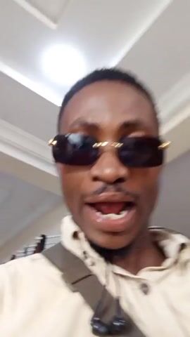 Here is a description of the video:

The video features a man wearing sunglasses and a light-colored shirt. He has a bag slung across his chest and appears to be indoors. He looks into the camera and moves his hands in the air. A light pink item, possibly clothing, is visible. The background shows a white ceiling with recessed lighting. A security camera is visible in one shot.