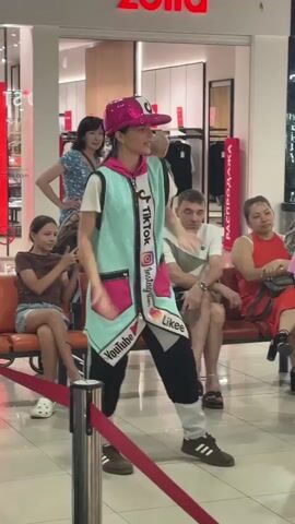 A young female dancer wearing a sparkly pink hat, a turquoise vest adorned with social media logos like TikTok, Instagram, and YouTube, and black pants with white stripes is performing in what appears to be a shopping mall. She is standing in front of a red velvet rope barrier, with several people seated on orange benches behind her, watching the performance. In the background, there are clothing displays and store interiors. The dancer is in motion, with her arms extended and body positioned as if in mid-dance move. The lighting suggests an indoor environment, likely during daylight hours due to the ambient light.