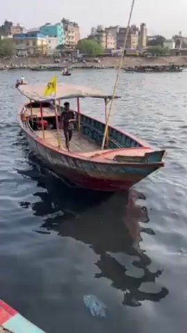 A medium shot captures a person pulling a boat toward the camera's vantage point. The individual is dressed in a dark t-shirt with light sleeves and dark athletic pants, as they are aboard a flat-bottomed boat painted with layers of red, blue, and green.  The boat has a white canopy and a flag, while a bamboo pole is stationed at its bow.  The boat is being pulled with a heavy rope. The river, stained a dark color, contains floating litter.  In the background, one observes a city skyline, partially obscured by trees. Other boats with passengers are sparsely scattered on the river.
