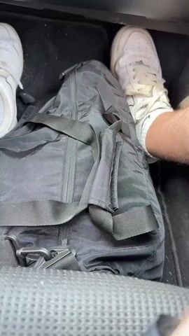 The video opens with a close-up shot of a car's front passenger footwell, showing a black duffel bag resting between two pairs of feet wearing white sneakers. The camera then pans up to reveal the car's dashboard and the view through the windshield. The car is driving on a paved road under a clear, bright blue sky. Green trees and fields line both sides of the road, and power lines are visible overhead. Another small, light-colored vehicle can be seen in the distance ahead on the road. A sticker is affixed to the top right corner of the windshield, but its text is illegible. The word "AIRBAG" is visible on the passenger side of the dashboard. The scene depicts a daytime drive through a rural or semi-rural area, likely Ezerovo, Bulgaria, as provided in the context.