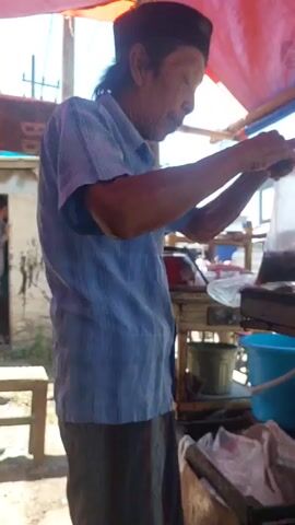 A side profile of an elderly East Asian man wearing a blue striped short-sleeved button-down shirt and a black cap is shown. He stands behind a food cart. His right hand holds a clear plastic bag containing a dark substance, and his left hand is near the bag. In the background, there are red and orange tarps, a blue bucket, and the suggestion of buildings and a street.